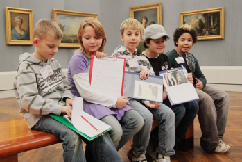 Fünf Kinder mit bunten Klemmbrettern sitzen auf einer Holzbank in einem Museum.