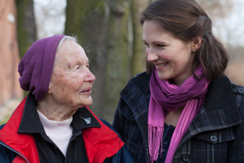 Eine ältere Dame und eine Ehrenamtliche lächeln sich an und sind im Wald unterwegs.